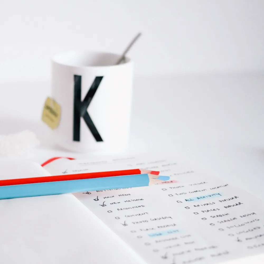 Close-up of a handwritten SEO checklist with red and blue pencils, placed beside a white mug labeled 'K'—symbolising content planning and optimisation.